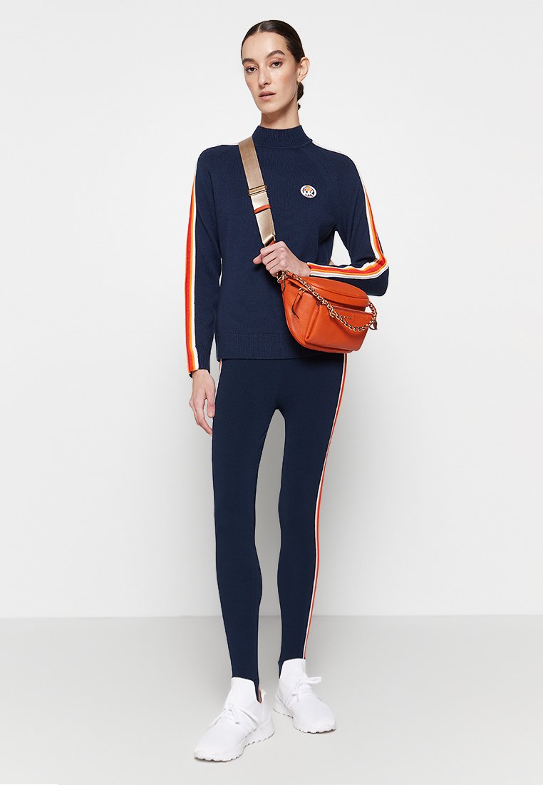 Vrouw in een marineblauw athleisure outfit met oranje en witte strepen, witte sneakers en een oranje schoudertas met een gouden ketting.