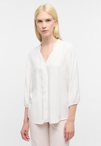 Weißes Bluse mit V-Ausschnitt, Rüschensaum und Knopfverschluss. Hat Dreiviertelärmel mit lockerer Passform und glatter Textur.