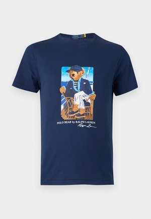 Homme debout portant un t-shirt bleu marine avec un motif d’ours, un pantalon blanc, une ceinture noire et des baskets beiges sur un fond blanc uni.