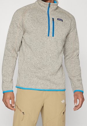 Mand iført beige fleece trøje med blå lynlåslomme på brystet og kant, kombineret med brune udendørs bukser med lynlåslommer.