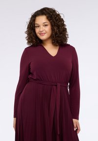 Bordowa sukienka z długimi rękawami i dekoltem w serek, z paskiem w talii i plisowanym dołem. Gładka, miękka tkanina o dopasowanym kroju góry.