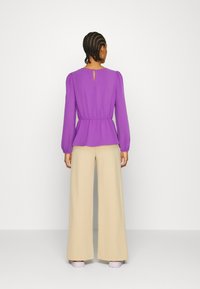 Blusa viola aderente con maniche a sbuffo e schiena con apertura a chiave, abbinata a pantaloni beige larghi e scarpe viola chiaro.