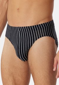 Homme portant un slip noir avec de fines rayures verticales blanches, montrant le torse et le haut des cuisses sur un fond uni.