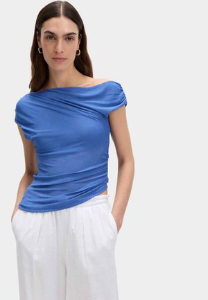 Femme aux longs cheveux foncés portant des boucles d'oreilles créoles dorées, un top asymétrique bleu décolleté épaule et un pantalon blanc taille haute, les mains dans les poches.