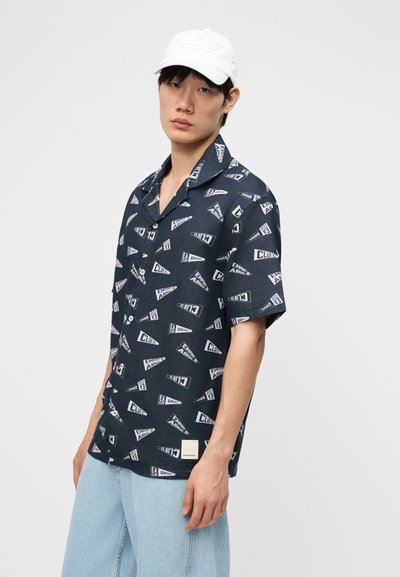 Νεαρός άνδρας που φοράει μπλε ναυτικό κοντομάνικο πουκάμισο με μοτίβα λευκού κειμένου και ανοιχτό μπλε τζιν, συνδυασμένο με λευκό καπέλο μπέιζμπολ.