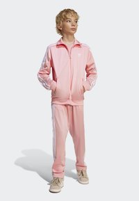 Rosa treningsdress laget av glatt stoff, med hvite striper langs ermene og buksene. Modell som har på seg beige joggesko med snøring.