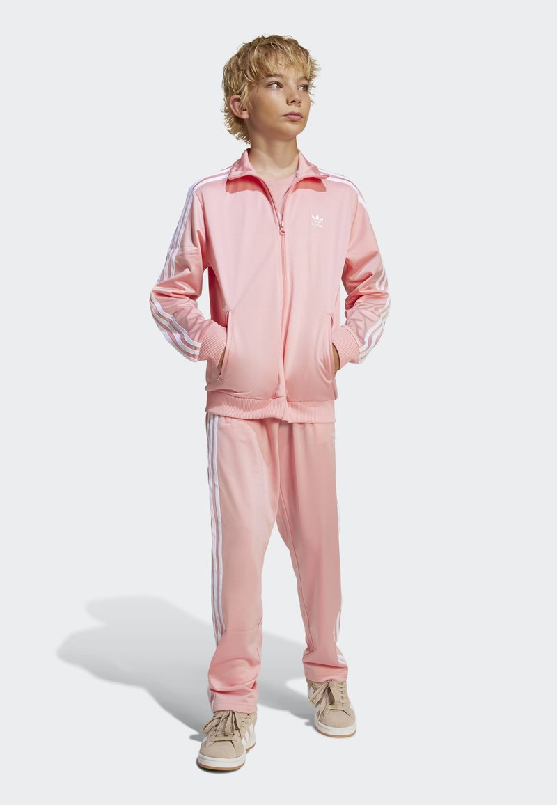 Rosa treningsdress laget av glatt stoff, med hvite striper langs ermene og buksene. Modell som har på seg beige joggesko med snøring.