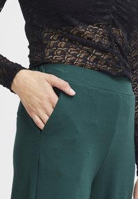 Černý krajkový top s dlouhými rukávy v kombinaci se zelenými, hladkými kalhotami s postranní kapsou. Detail ruky odpočívající v kapse.