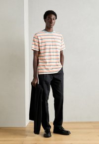 Stribet t-shirt i fersken, hvid og teal, parret med sorte bukser. Modellen holder en sort jakke, mens hun står på et trægulv.