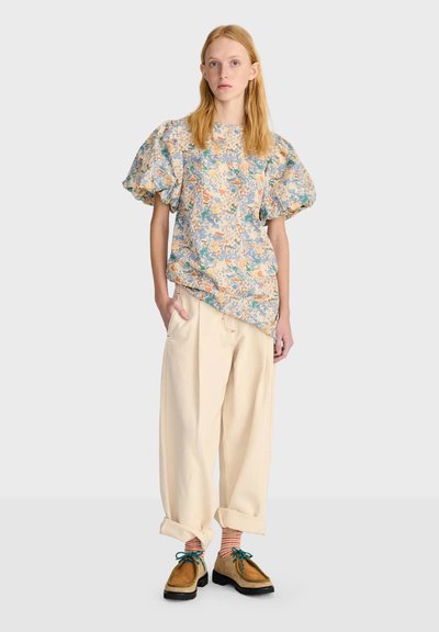 Jeune femme portant un chemisier à manches bouffantes fleuri, un pantalon beige ample retroussé aux chevilles, des chaussettes rayées et des chaussures marron à lacets sur un fond uni.