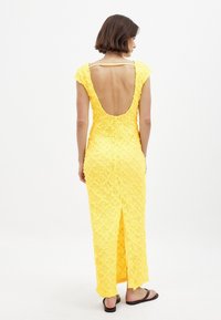 Robe en dentelle jaune avec un décolleté plongeant dans le dos, silhouette cintrée, motif floral et fente sur le côté, associée à des sandales noires.
