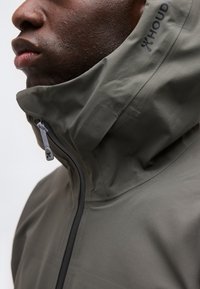 Gros plan sur une personne portant une veste à capuche vert olive, zippée jusqu'au menton, avec le nom de la marque "Houdini" visible sur la capuche.