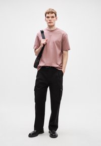 Růžové bavlněné tričko, volný střih, kombinované s černými cargo kalhotami s bočními kapsami. Model drží černou crossbody tašku. Černé boty.