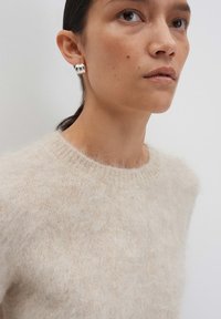 Pull en beige duveteux avec un col rond, doté d'une texture douce. Boucle d'oreille en argent avec un design texturé portée à l'oreille droite.