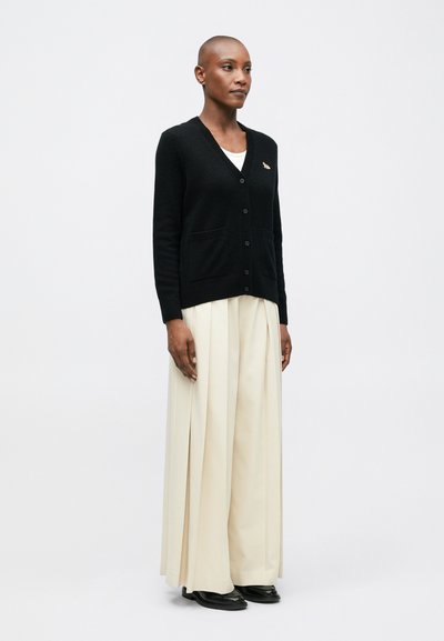 Cardigan negro con escote en V, cierre de botones, dos bolsillos frontales y un pequeño detalle bordado, combinado con una falda plisada de color crema.