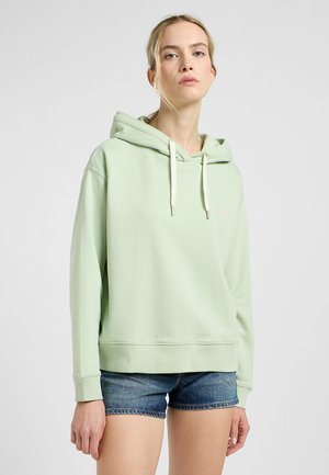 Felpa verde chiaro con cappuccio e cordino, caratterizzata da un fit rilassato e polsini a costine, esposta insieme a pantaloni shorts in denim.