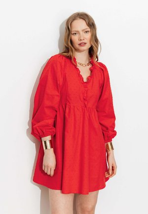 Femme portant une robe rouge à manches longues avec un col à volants, un collier chaîne en or et de larges bracelets manchettes dorés aux deux poignets.
