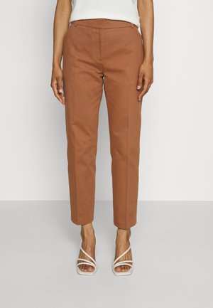 Woman wearing rust-colored straight-leg trousers and white strappy heeled sandals, standing on gray floor against white background.