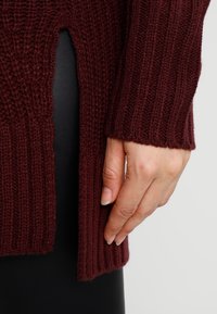 Burgundy ribbad stickad tröja med sidospalter, med en texturerad yta och långa ärmar. Hand som vilar på tyget visar detalj.