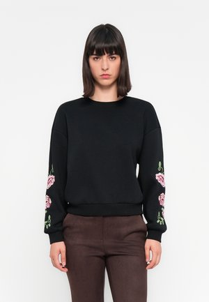 Donna con capelli scuri lisci, che indossa una felpa nera con ricami floreali rosa sulle maniche e pantaloni marroni, in piedi contro uno sfondo bianco.