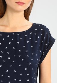 Marinblå topp med vitt ankarmönster, mjukt bomullsmaterial, rund halsringning, korta ärmar och avslappnad passform.