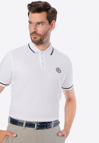 Bílá polo košile s černými detaily, na hrudi s logem. V kombinaci se světle zbarvenými kalhotami a tmavě modrým opaskem.