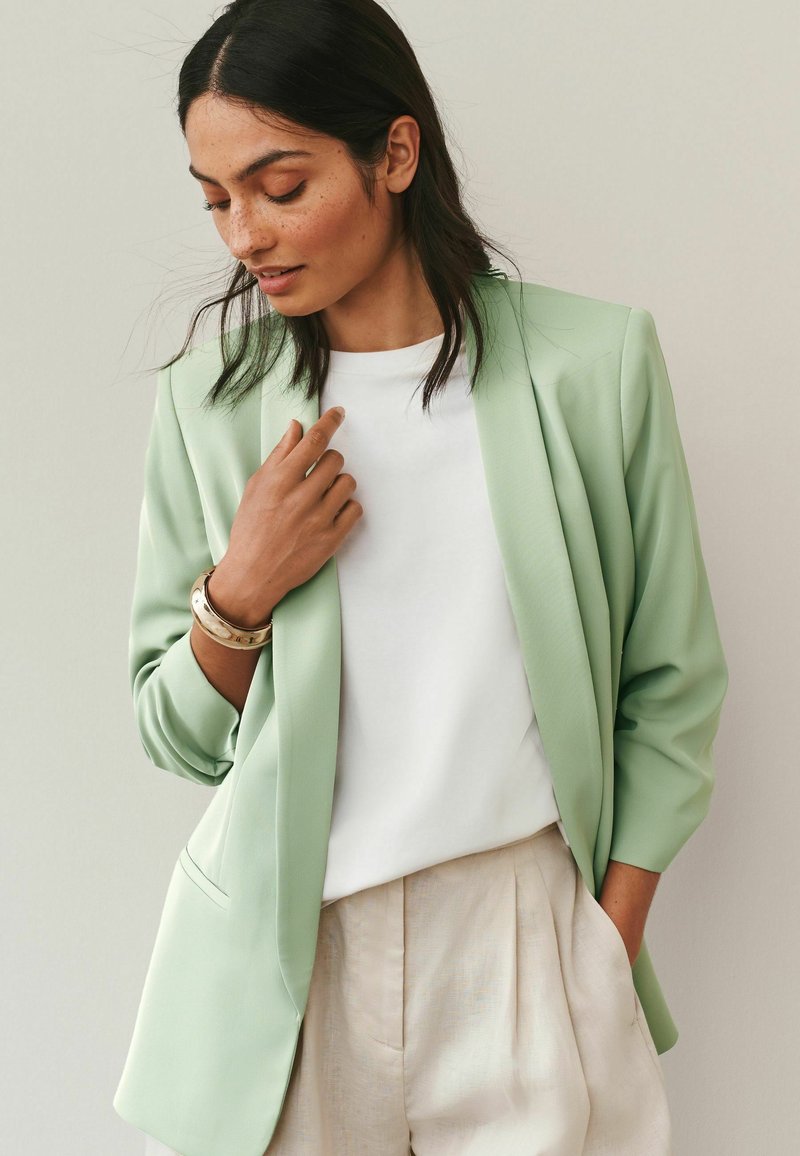 Femme aux cheveux foncés porte un blazer vert clair sur une chemise blanche et un pantalon plissé beige, accessoirisée d'un bracelet doré épais, regardant vers le bas.