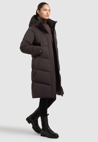 Abrigo acolchado largo de color marrón oscuro con capucha, diseño de líneas cosidas, dos bolsillos laterales y cierre con cremallera, combinado con pantalones negros ajustados y botines.