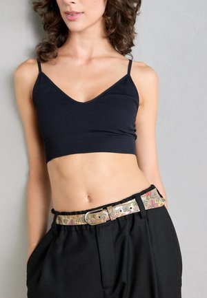 Woman wearing a black cropped tank top and high-waisted black pants with a floral patterned belt against a gray background.