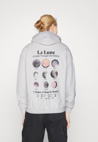 Ljust grå hoodie med en stor grafisk design på ryggen med himmelska kroppar och text. Ribbad muddar och midja; mjuk textur.