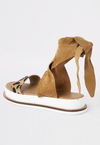 Tan-nahkatossut, joissa on leopardipainatettu hihna, avonaiset varpaat ja nilkkaketjut. Varustettu valkoisella alustalla, jossa on ruskea reunus.