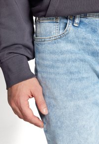Light blue denim jeans with a distressed texture, featuring a front pocket and contrast stitching, worn with a dark gray long-sleeve top.