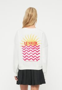 Femme blonde portant un sweat-shirt blanc avec un motif de soleil jaune et des vagues roses, texte rouge « LE SOLEIL », et une jupe plissée noire, vue de dos.