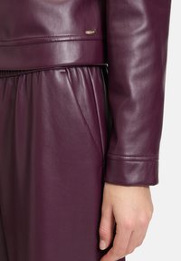 Głęboki purpurowy skórzany kurtka z dopasowanym mankietem i gładką teksturą, z lekko połyskującym wykończeniem oraz małym metalowym znaczkiem marki.