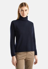 Kobieta z prostymi blond włosami nosi granatowy sweter z golfem oraz beżowe spodnie, stojąc na tle jasnego, jednolitego tła.