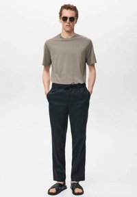 Grijze kortekous t-shirt, zwarte elastische taillebroek en zwarte sandalen. Model draagt een zonnebril, met handen in de zakken. Eenvoudig en casual ontwerp.