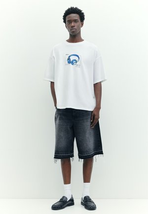 Junger Mann trägt ein weißes übergroßes T-Shirt mit einem blauen Kopfhörer-Aufdruck, dunkle ausgefranste Jeansshorts, weiße Socken und schwarze Loafers vor weißem Hintergrund.