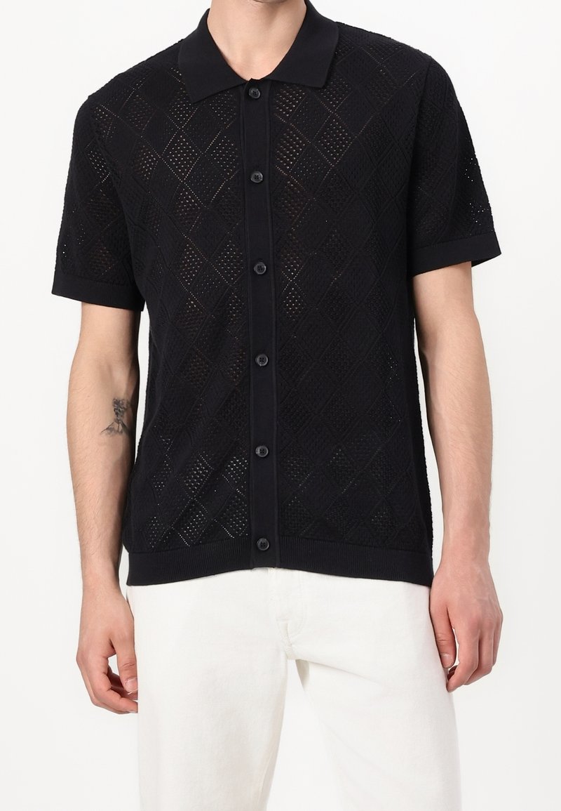 Polo noir en maille à manches courtes avec motif en maille losange, fermeture à boutons et col contrastant. Texture lisse, coupe ajustée.