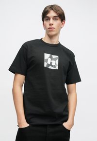 Μαύρο βαμβακερό μπλουζάκι με κεντρικό λογότυπο σε σχήμα αστεριού σε γκρι απόχρωση. Ο σχεδιασμός διαθέτει στρογγυλή λαιμόκοψη και κοντά μανίκια.