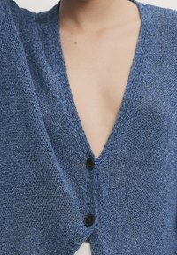 Blå stickad kofta med djup V-ringning, som har två svarta knappar och ett texturerat tyg som framhäver vävdetaljer och mjukhet.