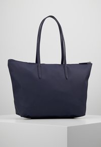 Sac cabas bleu marine avec deux longues poignées, fabriqué en tissu résistant. Le design présente une surface lisse et une fermeture éclair sur le dessus.