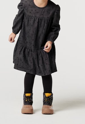 Kleinkind trägt ein schwarzes Kleid mit Sternenmuster, schwarze Leggings und braune Winterstiefel mit schwarzen Schnürsenkeln und gelbem Innenfutter.