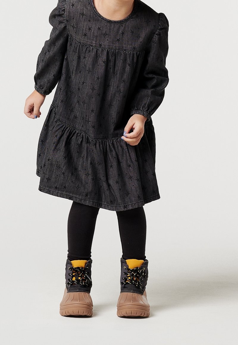 Tout-petit portant une robe noire à motifs d'étoiles, des leggings noirs et des bottes d'hiver couleur sable avec des lacets noirs et une doublure intérieure jaune.