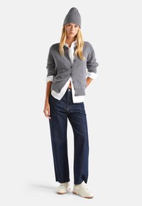 Cárdigan gris de punto, camisa blanca de botones, jeans de pierna ancha azul oscuro y zapatillas crema. El modelo lleva un gorro gris. Diseño sencillo, textura suave.