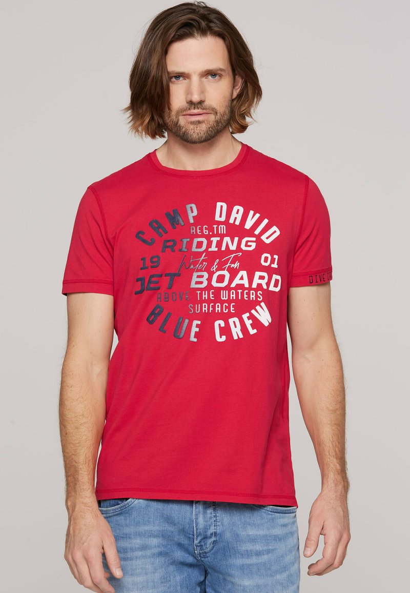 T-shirt in cotone rosso con grafiche testuali circolari in bianco e grigio, scollatura rotonda e maniche corte. Il design include dettagli del marchio.