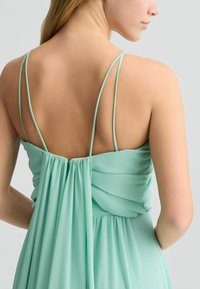 Abito di un delicato verde menta con spalline sottili e schiena arricciata. Presenta pieghe morbide, tessuto fluido e una piccola chiusura dorata al collo.
