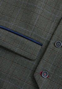 Kostkovaný vlněný blazer v tmavě zelené a šedé barvě, s modře lemovanou kapsou a černým knoflíkem s červeným detailem. Texturovaná látka se strukturovaným finišem.