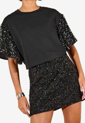 Žena nosící černý top s flitrovými rukávy a odpovídající černou mini sukni s flitry, doplněno stříbrným náramkem.