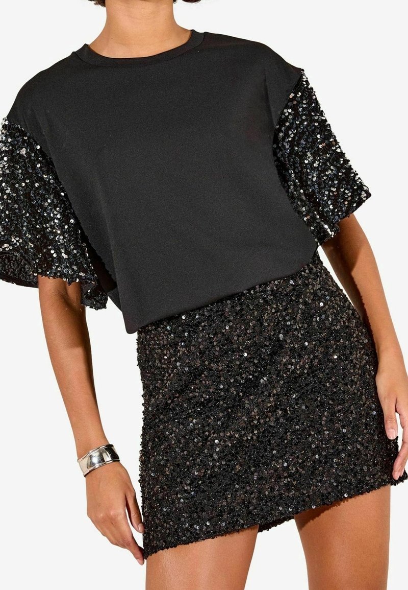 Femme portant un top noir avec des manches à sequins et une mini-jupe noire à sequins assortie, accessoirisée d'un bracelet argenté.