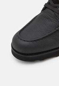 Chaussure en cuir noir avec un bout arrondi, des détails cousus et une surface texturée. La semelle est épaisse et légèrement surélevée.